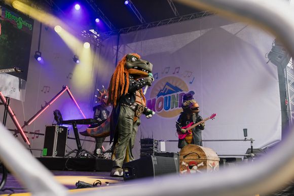 Dinosaurierkostüme spielen auf einer Bühne mit bunten Lichtern, ein Musiker singt ins Mikrofon.