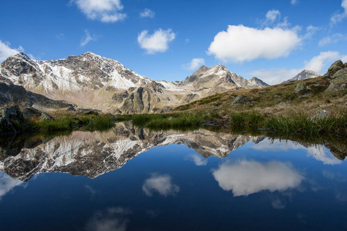 Klarer Bergsee reflektiert umliegende schneebedeckte Gipfel; Uferbereich mit Gras und Felsen, blauer Himmel mit einzelnen Wolken.