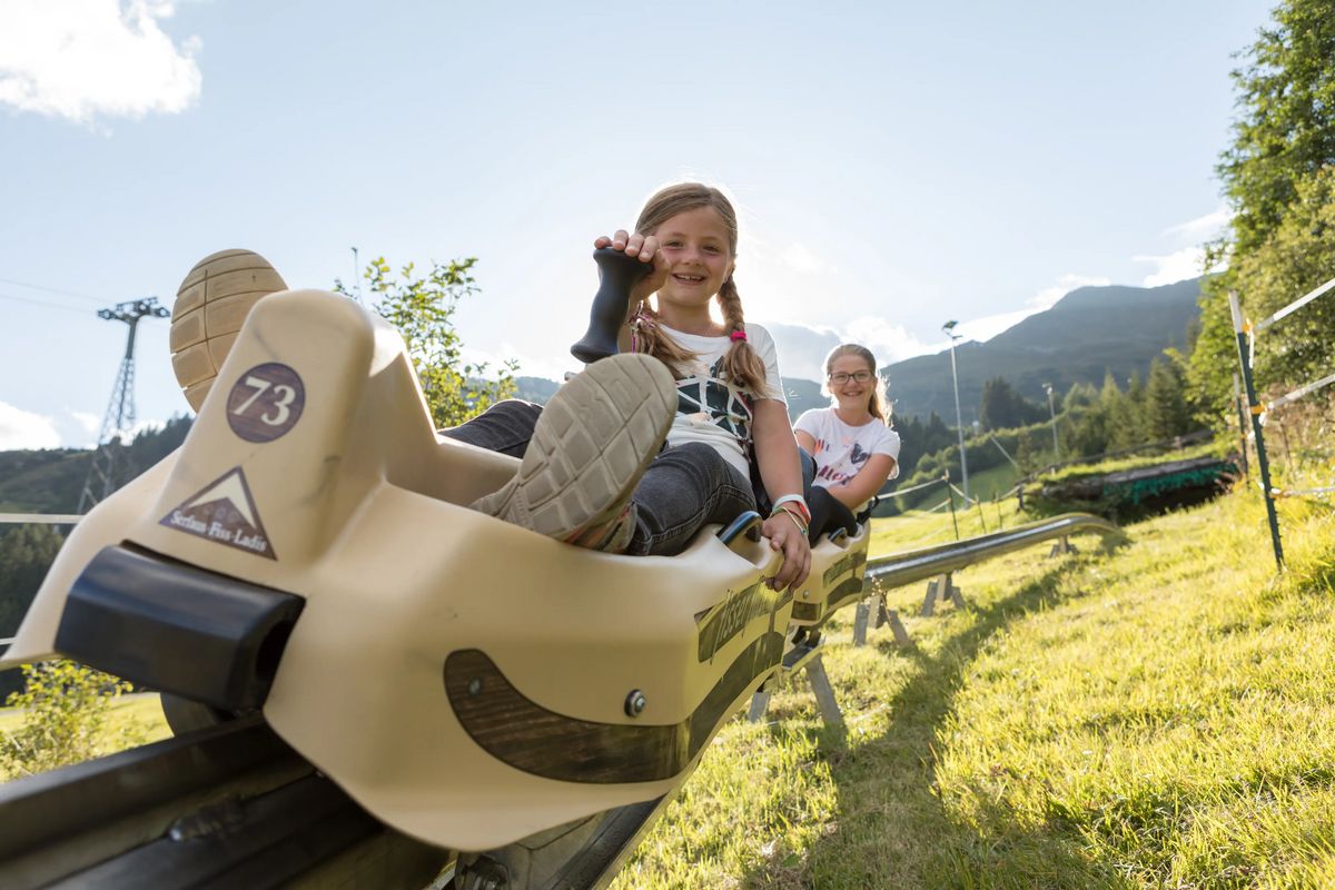 Zwei Kinder sitzen in einer Schienen-Rodelbahn und fahren bergab, dahinter Wiesen, Bäume und Berge bei sonnigem Wetter.