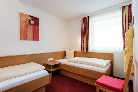 Zweibettzimmer mit Holzmöbeln, rotem Teppich, Fenster mit Vorhängen und schlichtem Wandbild.