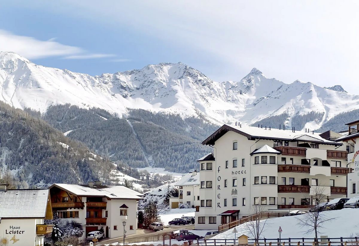 Hotelgebäude in verschneiter Ortschaft mit Blick auf hohe, schneebedeckte Berge im Hintergrund.