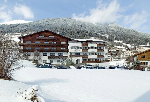 Panorama eines Hotels vor verschneiten Bergen, mit Parkplätzen und umliegenden Winterlandschaften bei klarem Wetter.