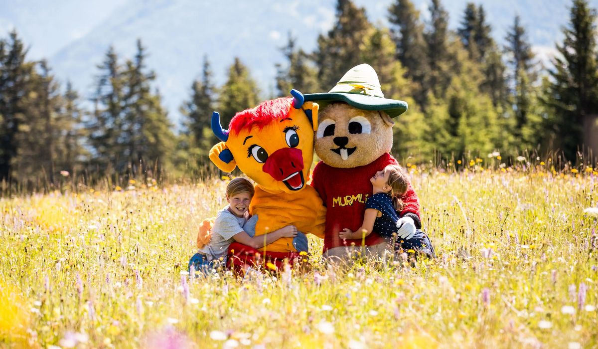 Zwei Kinder umarmen zwei große Maskottchen in Tierkostümen auf einer bunten Blumenwiese, im Hintergrund Nadelbäume und Berge.