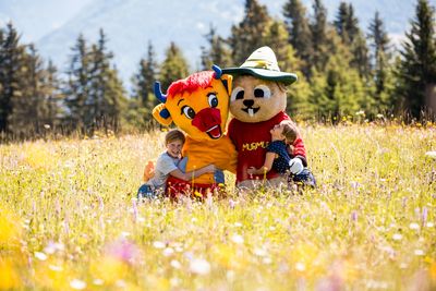 Zwei Kinder umarmen zwei große Maskottchen in Tierkostümen auf einer bunten Blumenwiese, im Hintergrund Nadelbäume und Berge.